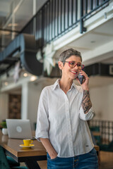 One senior woman with tattoo on left hand standing at cafe and talking on mobile phone, modern...