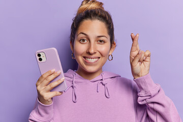 Positive young European woman holds cellphone winking eye and crossing her fingers in hopeful...