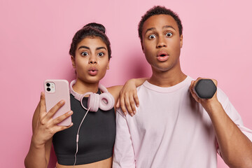 Sporty lifestyle concept. Shocked woman and man train in gym with dumbbells hold smartphone stare stupefied at camera stand closely to each other against pink background hear something shocking