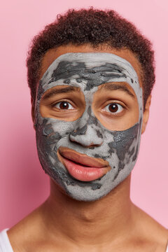 Face care concept. Puzzled African man wwith curly hair applies beauty clay mask purses lips concentrated at camera undergoes well being procedures focused at camera isolated over pink background.