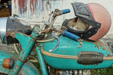 part of an old rusty iron broken heavy green dirty made in the ussr motorcycle with a dirty plastic red hardhat on the tank on the street at daytime
