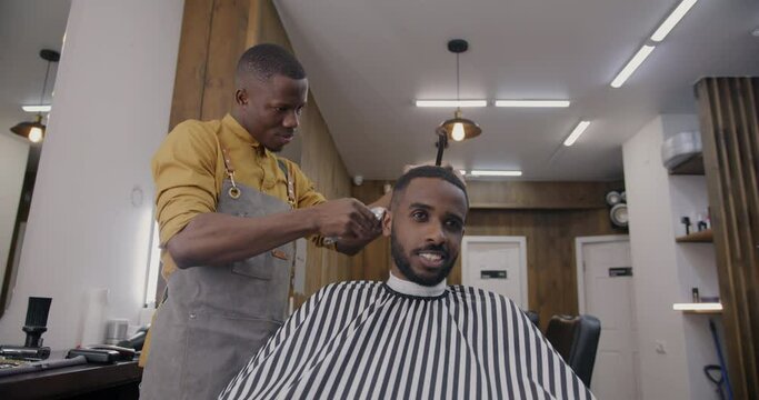 Joyful guy talking and smiling while barber shaving hair with machine in barbershop. Positive emotion and beauty salon activity concept.