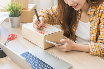 Online seller store, owner small business entrepreneur asian young woman packing product in box, using smartphone, mobile check purchase order from web prepare parcel sent delivery company to customer
