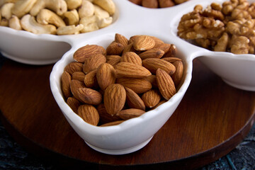 On a wooden stand there is a beautiful white plate in the shape of petals with nuts laid out in it. Different types of delicious nutritious nuts on a plate for a healthy snack