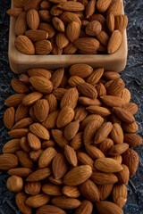 Delicious roasted almonds close-up on a gray textured background are scattered from a square-shaped wooden plate. Raw peeled almonds stacked in a heap on the table, top view
