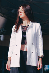 
beautiful young woman in a white jacket and black trousers with an open belly and a tattoo in the city against the backdrop of glass buildings