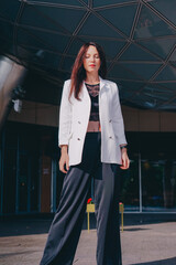 
beautiful young woman in a white jacket and black trousers with an open belly and a tattoo in the city against the backdrop of glass buildings