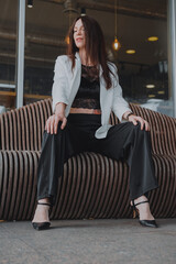
beautiful young woman in a white jacket and black trousers with an open belly and a tattoo in the city against the backdrop of glass buildings