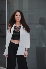 
beautiful young woman in a white jacket and black trousers with an open belly and a tattoo in the city against the backdrop of glass buildings