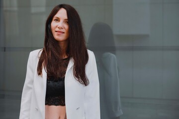 
beautiful young woman in a white jacket and black trousers with an open belly and a tattoo in the city against the backdrop of glass buildings