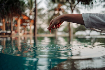 An elegant woman's hand touches the surface of the turquoise water, creating ripples, symbolizing...