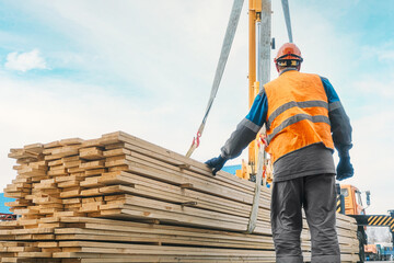 A slinger unloads wooden planks outdoors on a summer day. A worker in a hard hat and high-visibility vest stacks lumber. Industrial background with copy space.