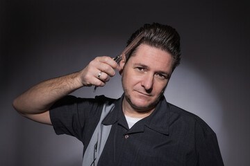 Horizontal studio portrait of a 40 year old man in rockabilly style combing his toupee with a steel...
