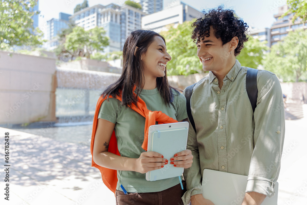 Wall mural two smiling multiracial student holding digital tablet, laughing together in university campus. happ