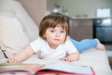 A caucasian little girl of two years with blue eyes in a white T-shirt lies on a sofa with a book.