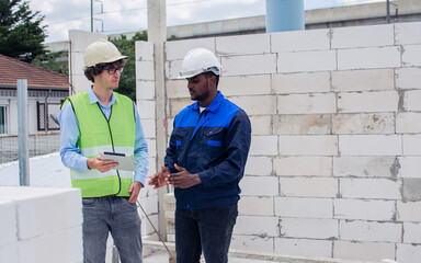 Fototapeta na wymiar Two diversity male engineers team working, inspecting outdoor at construction site, wearing hard hats for safety, talking, discussing, using tablet to plan for building. Career, Industry Concept.