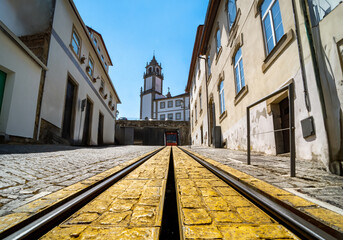 Yellow tram track with cobblestones and metal rails from the top of the sloping street where you can see the cathedral of Viseu at the top of the street and the red wagon in the garage - obrazy, fototapety, plakaty