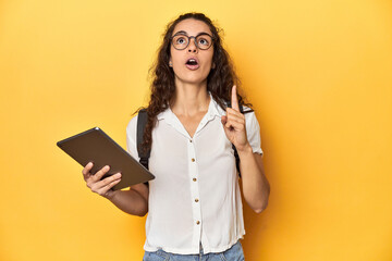 Student holding a tablet, glasses, backpack on, pointing upside with opened mouth.