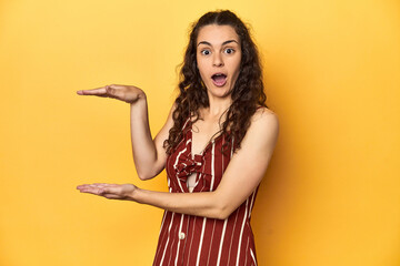 Young Caucasian woman, yellow studio background, shocked and amazed holding a copy space between hands.