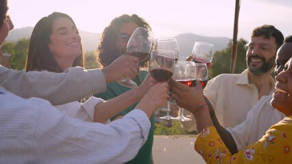 Gathering of multi-ethnic people celebrating rooftop party toasting with red wine at sunset. Group of young friends have fun laughing on terrace on summer day. Socializing and festivities millennial. 