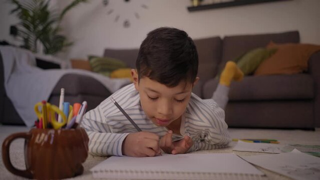 Little Latino boy draws quietly lying in living room at home. Concentrated child doing school homework at weekend. Concept of kids activities on vacations and family free time in childhood. 