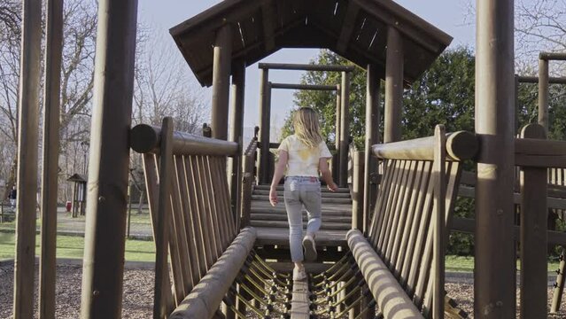 Active Caucasian little girl plays on playground bridge on sunny day. Blonde girl turns to camera in wooden house and poses happy and cheerful. Childhood fun outdoor with family enjoying leisure.