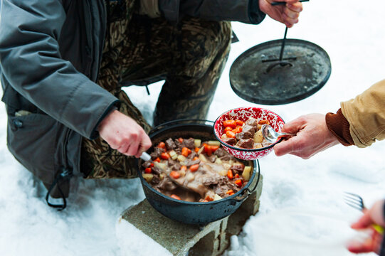 A man serves hot venison stew onto a dish from a cast iron pot in the snow