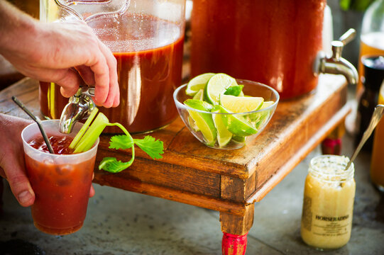 A hand pours bloody mary mix from a glass dispenser into a cup with celery, with limes and horseradish nearby