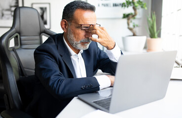 Unhappy sad tired mature european man in suit work on laptop, has stress, migraine in office interior