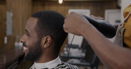 Joyful client smiling while barber brushing and drying his hair with electric dryer in barbershop. Modern appliance and beauty salon concept.