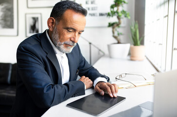 Fototapeta na wymiar Serious handsome mature european businessman in suit work on tablet with empty screen in light office