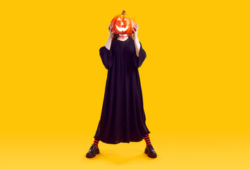 Unrecognizable woman hides her face behind glowing Halloween pumpkin with scary carved face. Full length woman in long black dress isolated on orange background. Halloween concept. Web banner.