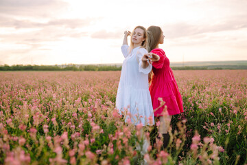 Two woman in stylish summer dresses feeling free in the field with flowers in sunshine. Nature, vacation, relax and lifestyle. Summer landscape.