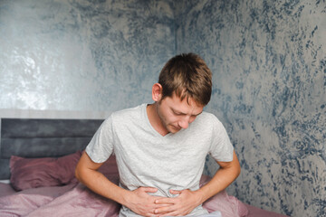 Young Caucasian man suffering from stomach pain while sitting on the couch at home and touching his...
