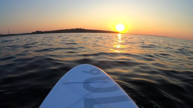 White SUP board with yellow oar on surface of sea at sunset dawn. Person swims on stand-up paddleboard in ocean near shore at sunrise sunset. POV. Wide below angle. Active lifestyle sport recreation