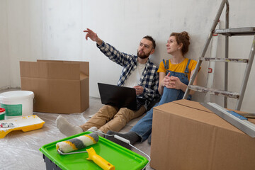 Couple sit on floor in new apartment during renovation process with laptop. Planning for furniture...