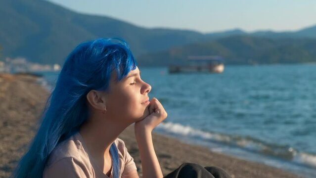 Fashioned teen with dreaming mood. A pretty blue hair girl relax on the shore by turquoise sea on tropical bay in the day light.