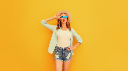 Summer portrait of beautiful young woman blowing her lips sending sweet air kiss wearing straw round hat on yellow background