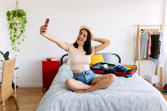 Happy pretty woman taking selfie with phone while preparing travel suitcase. Smiling traveler female posing for self portrait sitting on bed before going on holidays.
