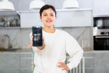 Cheerful young woman in casual white clothing standing in cozy home kitchen extending hand with phone, showing blank screen..