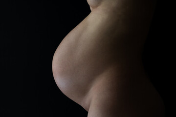 Midsection of unrecognizable standing naked mother with very round pregnant baby bump. Final month of pregnancy - week 36. Side view. Black background.