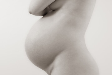 Middle part of unrecognizable standing naked mother with very round pregnant baby bump. Final month of pregnancy - week 36. Side view. White background. Bright shot. Sepia.