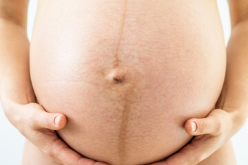 Closeup of very round pregnant baby bump held by hands of naked standing mother gently holding. Front view. White background. Bright shot.