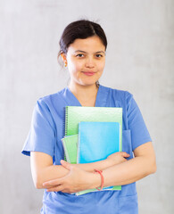 Obraz premium Portrait of smiling young female doctor in blue uniform standing with notebooks indoors on gray background..