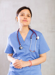 Friendly young female professional doctor in blue scrub with phonendoscope around neck posing against gray studio background, looking at camera with smile..