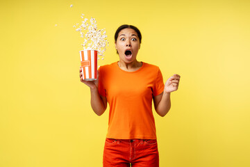 Excited African American woman holding bucket with popcorn, eating snacks watching movie with open  mouth in isolated on yellow  background. Cinema time, advertisement concept