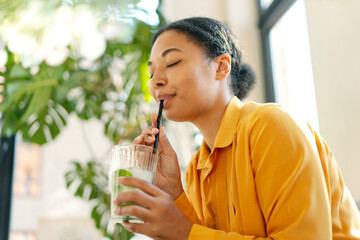Portrait of smiling beautiful African American woman drinking drinking fresh tasty lemonade or cocktail sitting and relaxing in modern caffe. Summer concept - obrazy, fototapety, plakaty