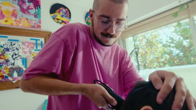 Young bald man with beard and mustache shaving hair of his girlfriend with electric razor, giving a haircut at home. Medium handheld shot