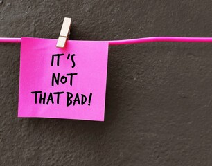 Pink note stick on dark wall with handwritten text IT’S NOT THAT BAD!, positive self talk or...