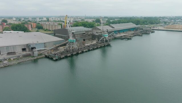 Aerial establishing view of port cranes and empty loading docks at Port Of Liepaja (Latvia), Liepaja city in the background, overcast summer day, wide slow ascending drone shot moving backward